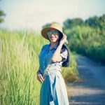 Stylish woman in a straw hat smiles amidst lush greenery on a sunny day.